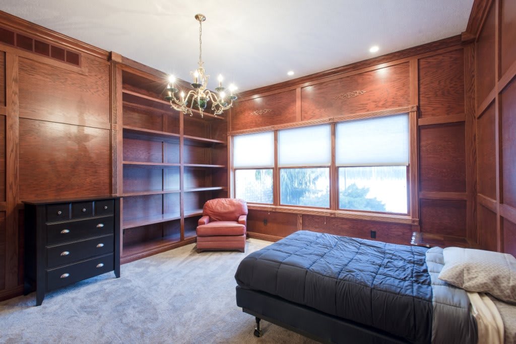 A cozy bedroom with wood paneling, featuring built-in bookshelves, a comfortable armchair, a bed, and a chandelier providing elegant lighting.