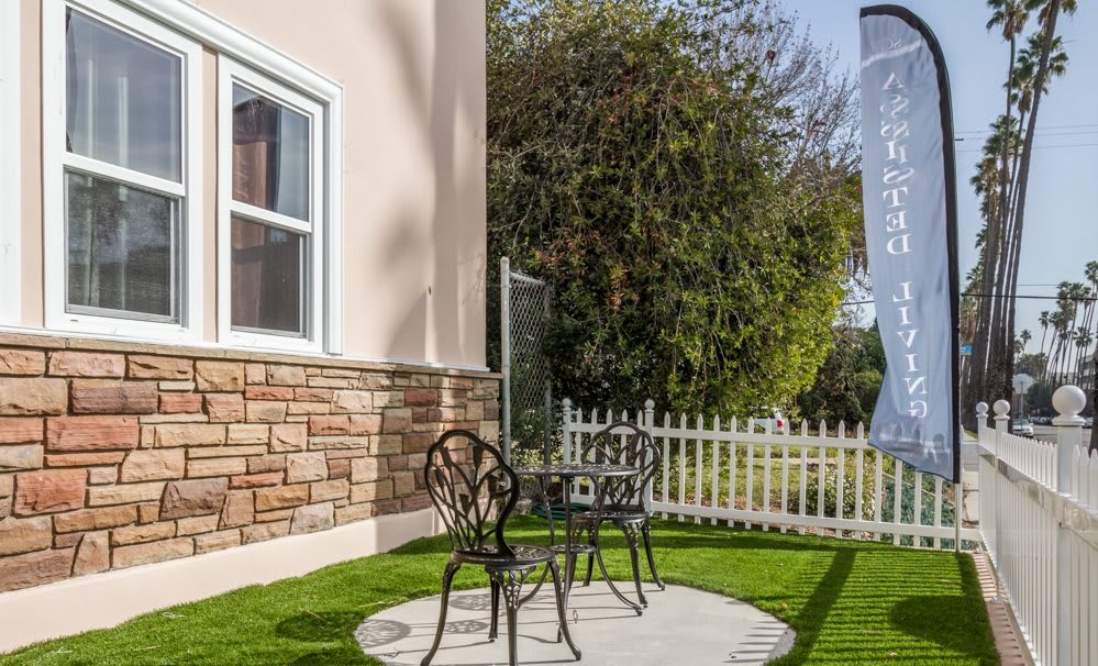 A cozy outdoor patio area with a small table and two chairs on artificial grass, surrounded by a white picket fence.