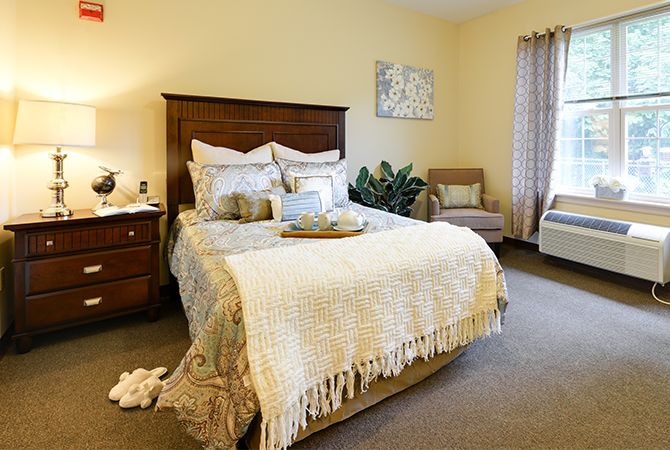 A cozy bedroom with a well-decorated bed, nightstand, and a comfortable chair by the window.