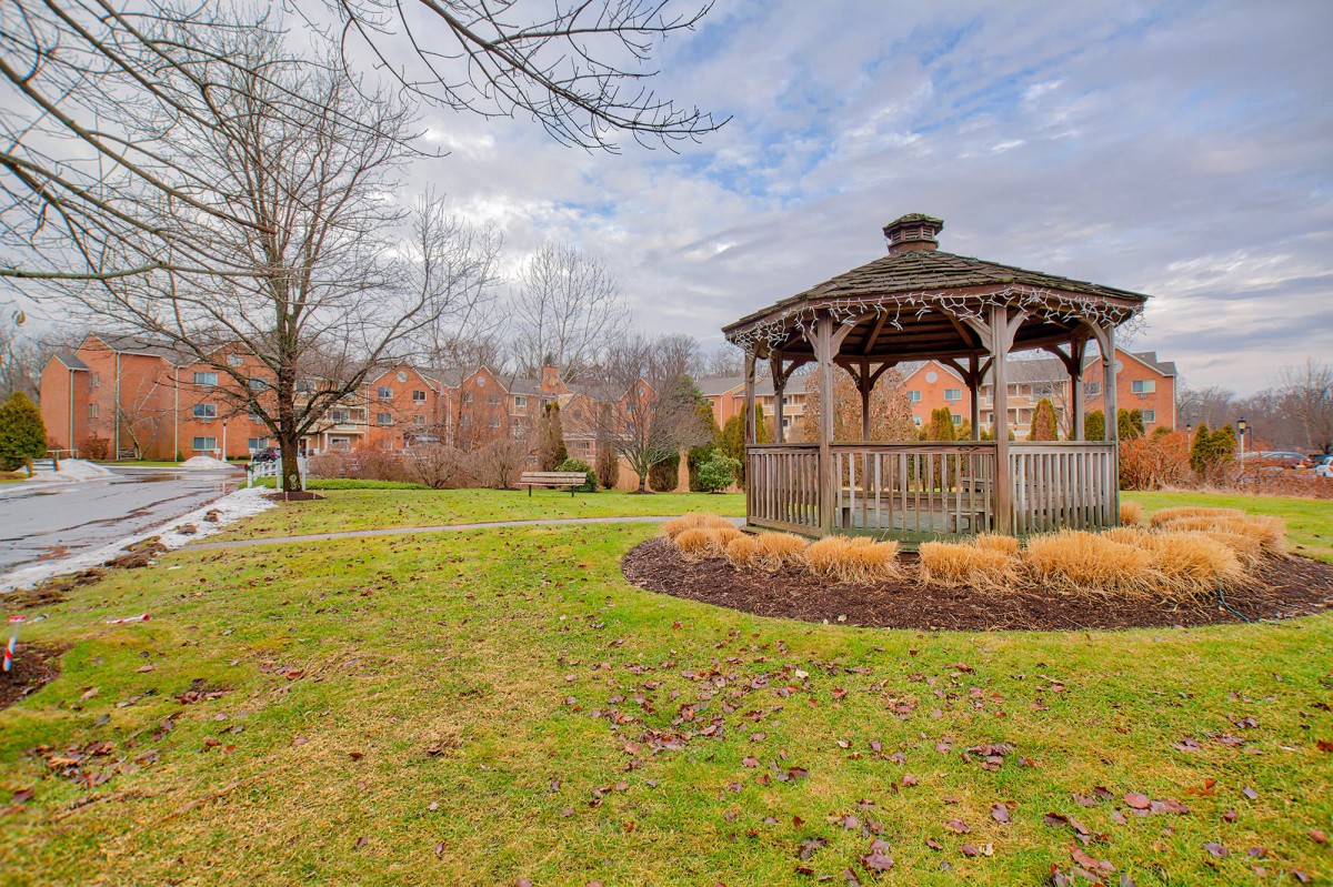 A serene park scene featuring a wooden gazebo surrounded by grass and trees under a cloudy sky.