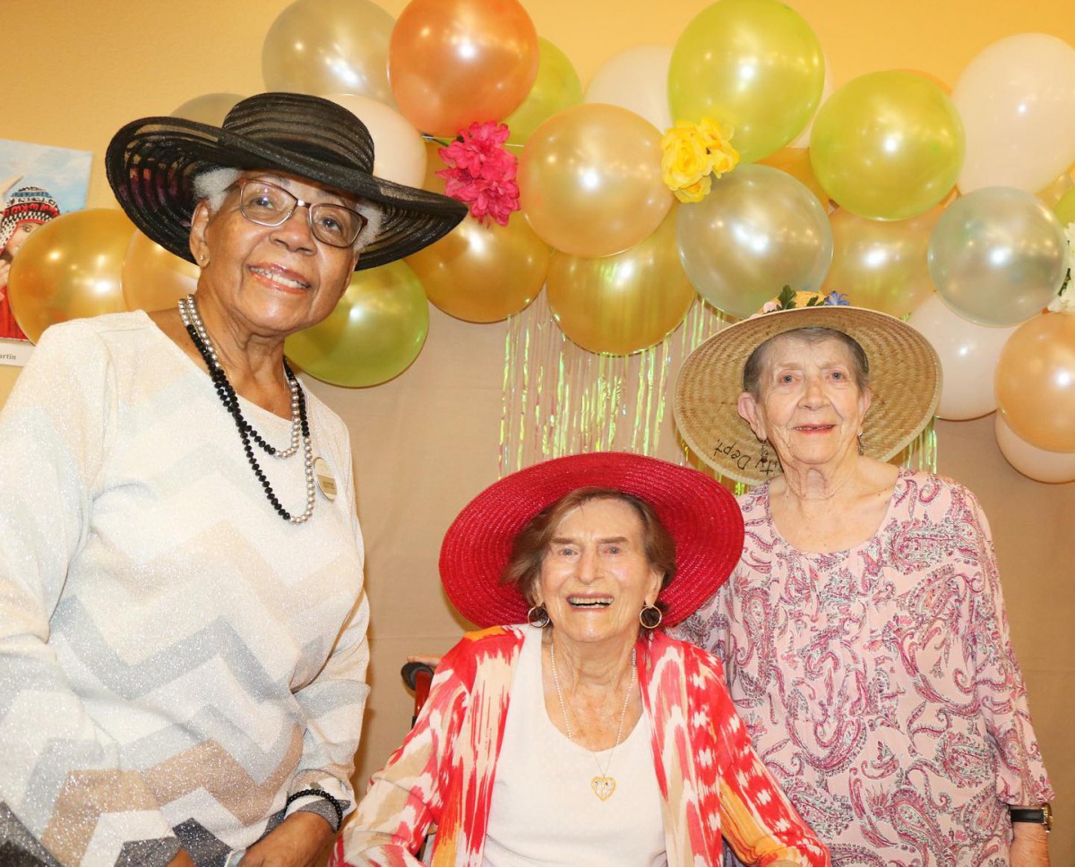 Three elderly women wearing hats smile for a group photo at a celebration with balloons in the background.