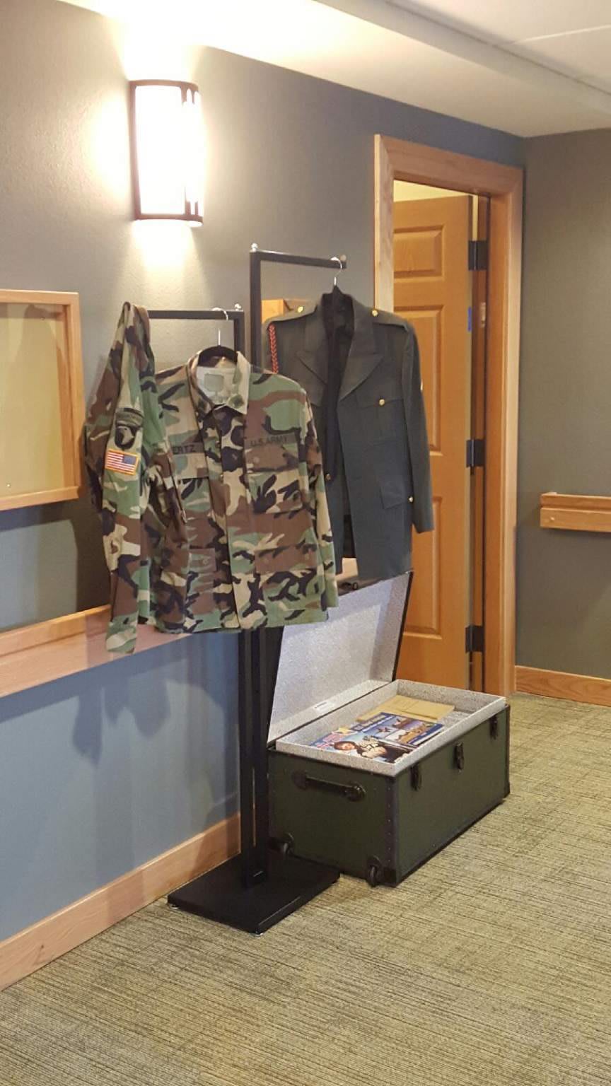 A display of military uniforms, featuring a camouflage jacket and a formal jacket hanging above an open trunk.