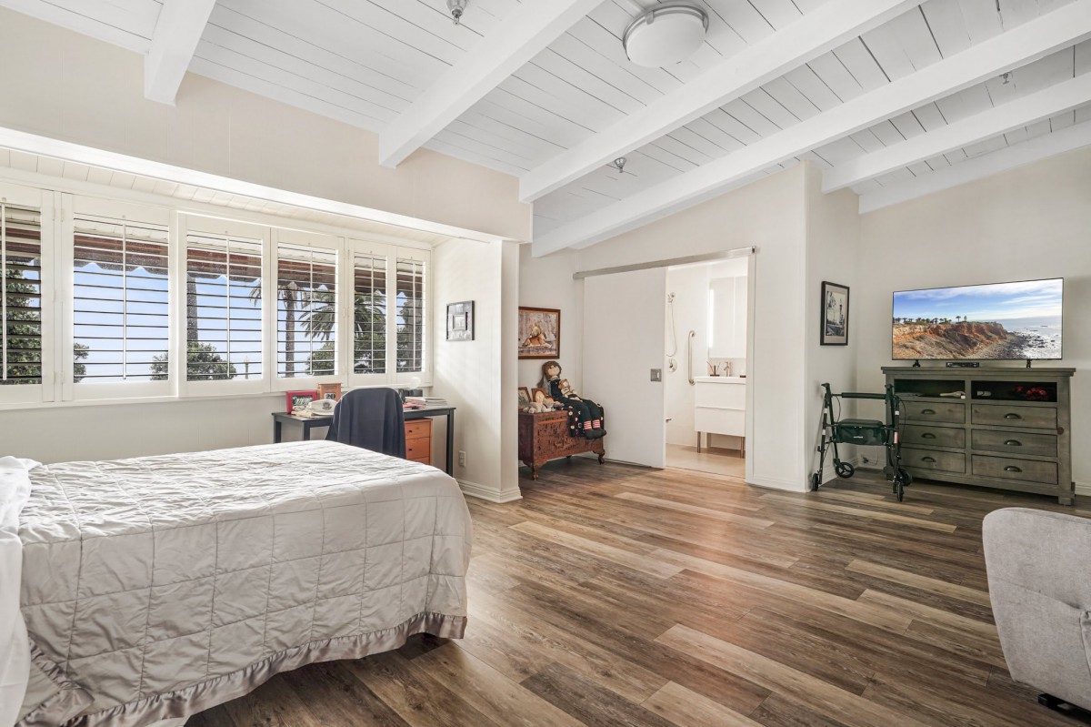 A cozy bedroom with wood flooring, ceiling beams, and ample natural light.