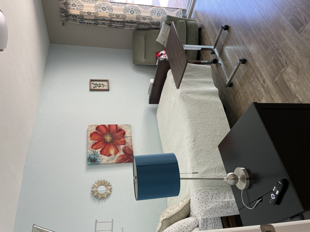A neatly arranged room featuring a single bed, blue lamp on a side table, and a flower painting on the wall.