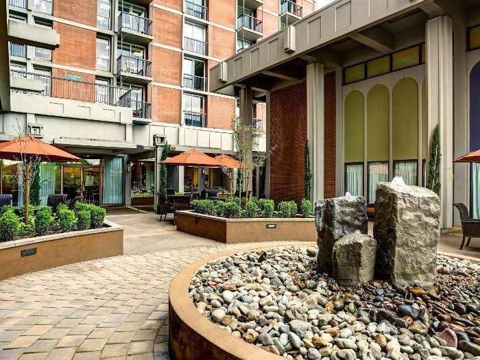 A modern courtyard featuring outdoor seating, landscaped plants, and a small fountain surrounded by buildings.