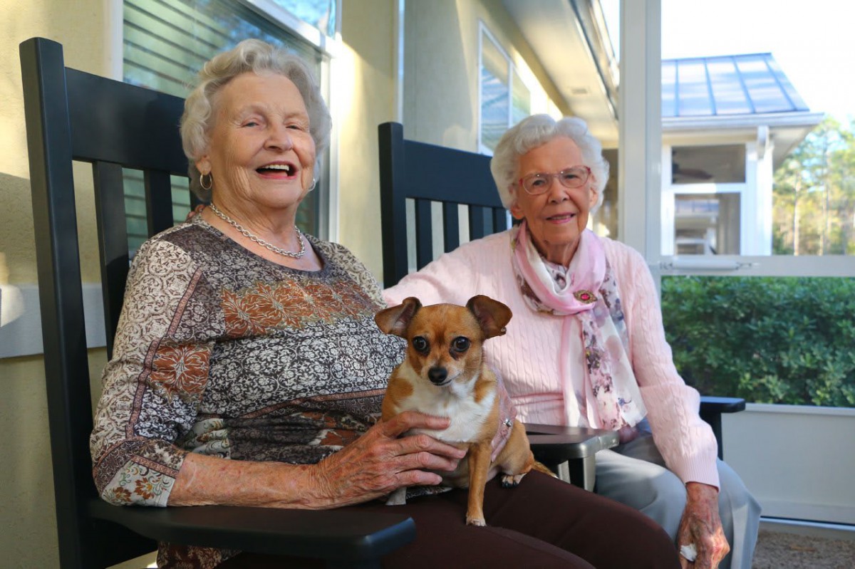 Two elderly women sitting on rocking chairs with a dog on a porch, smiling and enjoying the outdoors.