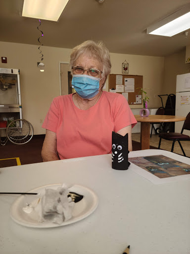 A senior woman wearing a mask sits at a table, engaged in a social activity.