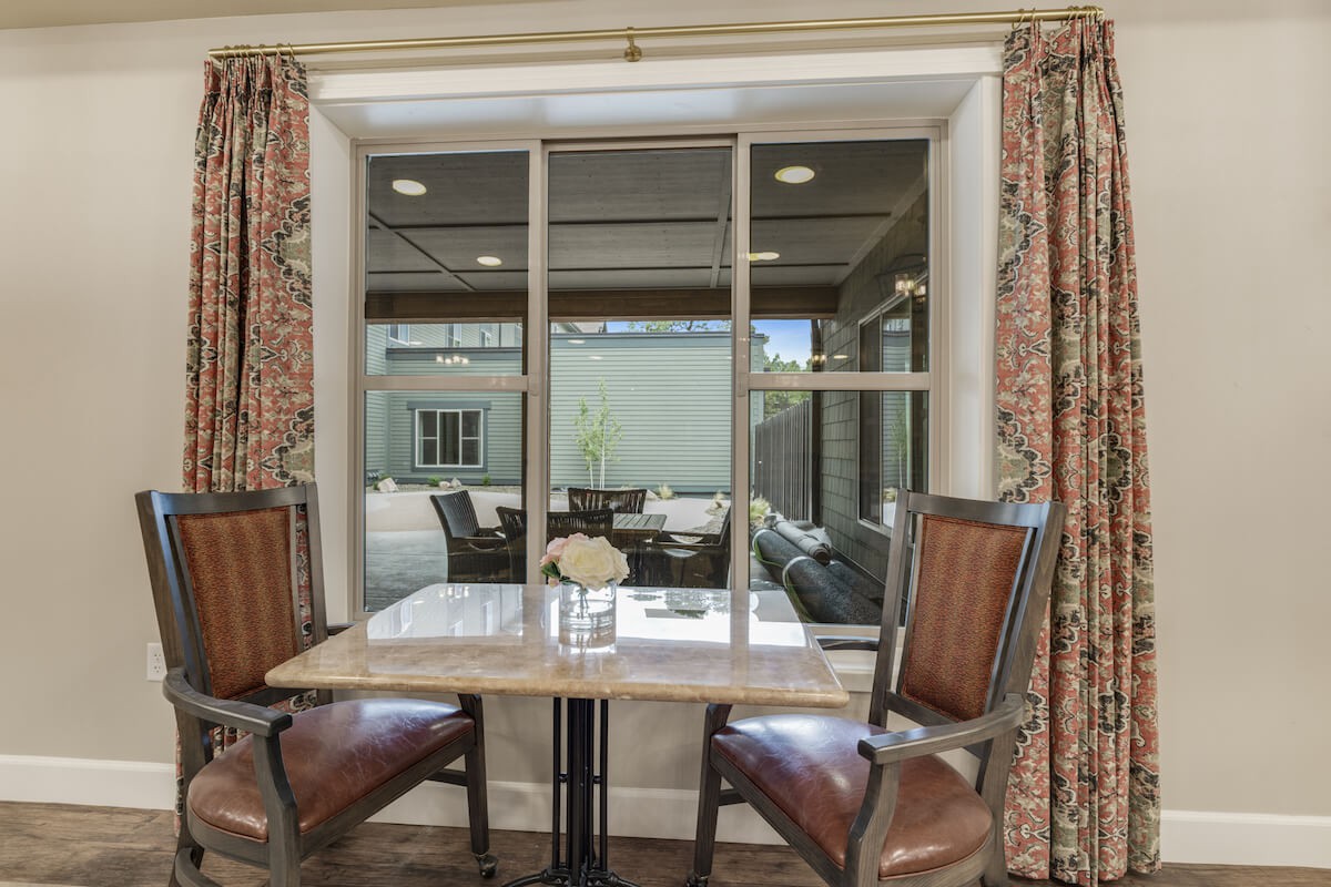 A cozy indoor dining area with a table and two chairs, featuring a large window and decorative curtains.