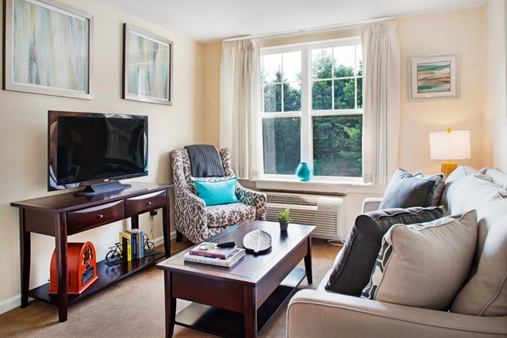 A cozy living room with a sofa, armchair, and coffee table, illuminated by natural light from a large window.