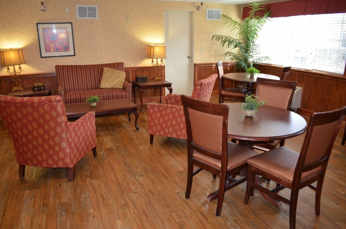 A cozy living room setting with wooden flooring, featuring a sofa, armchairs, dining tables, and an indoor plant.
