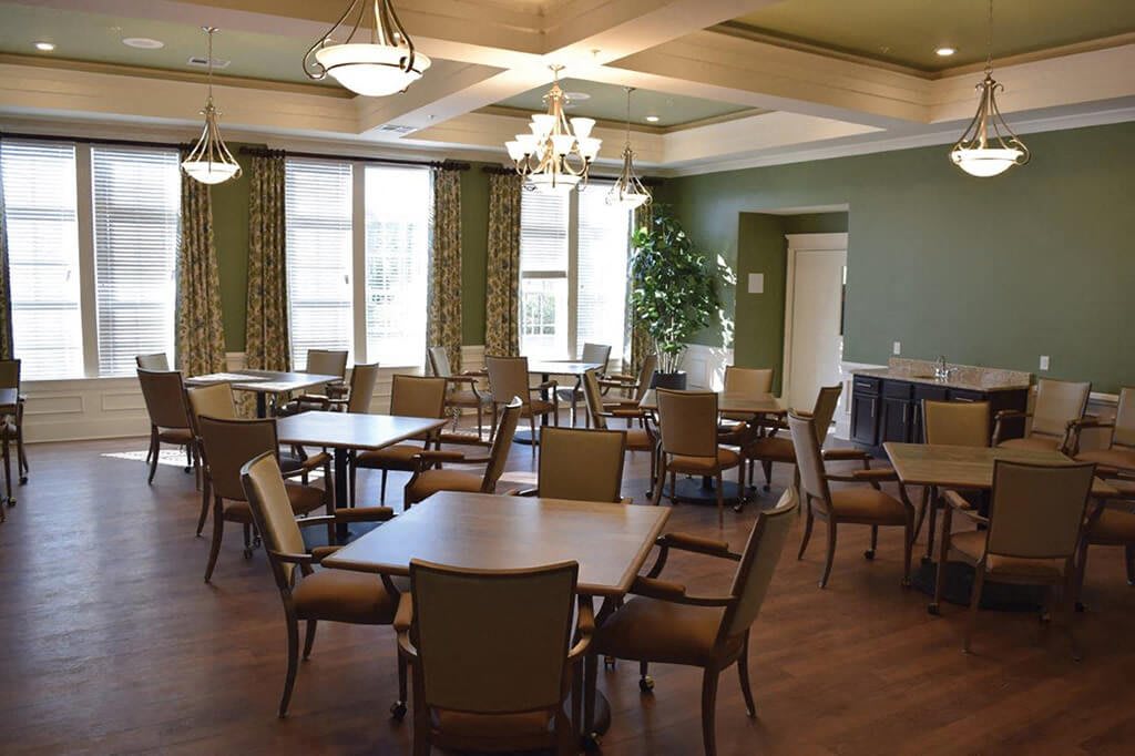 A well-lit dining room with neatly arranged tables and chairs, featuring large windows and elegant chandeliers.