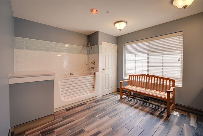 A minimalist bathroom with a bathtub, hardwood floor, and a wooden bench by the window.