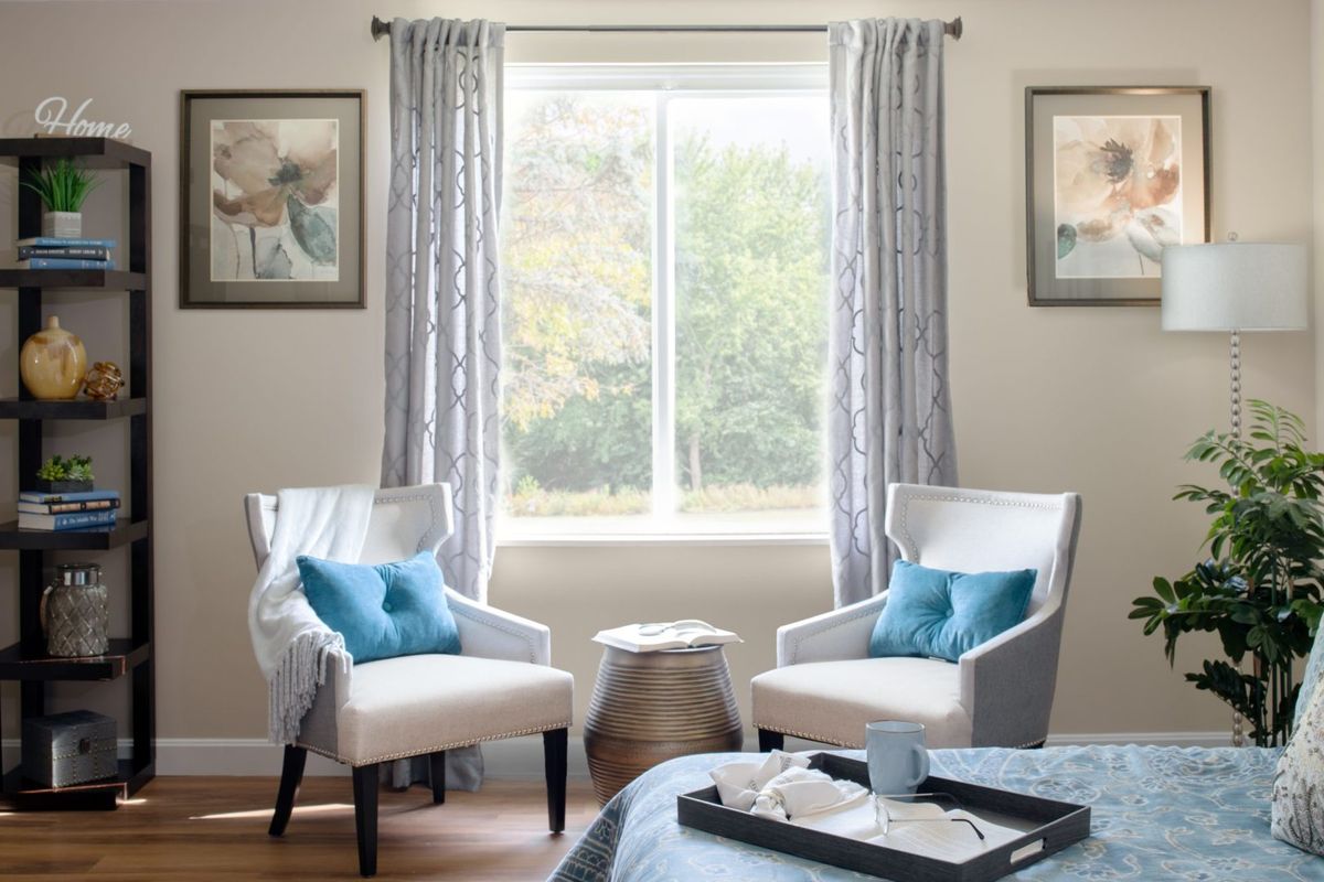A cozy living room corner featuring two white chairs with blue pillows, a small table, and natural light streaming in through the window.