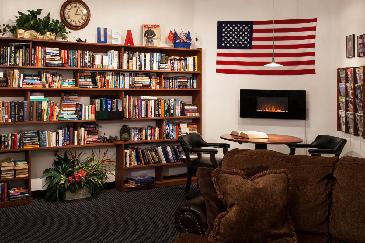 A cozy reading room featuring a large bookshelf filled with books, an American flag on the wall, and a comfortable armchair near a fireplace.