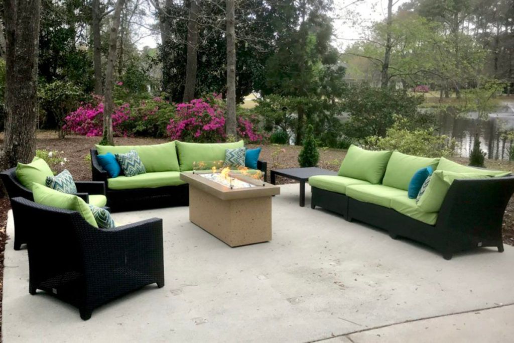 A cozy outdoor patio setup with green-cushioned seating and a central fire pit surrounded by blooming garden plants.