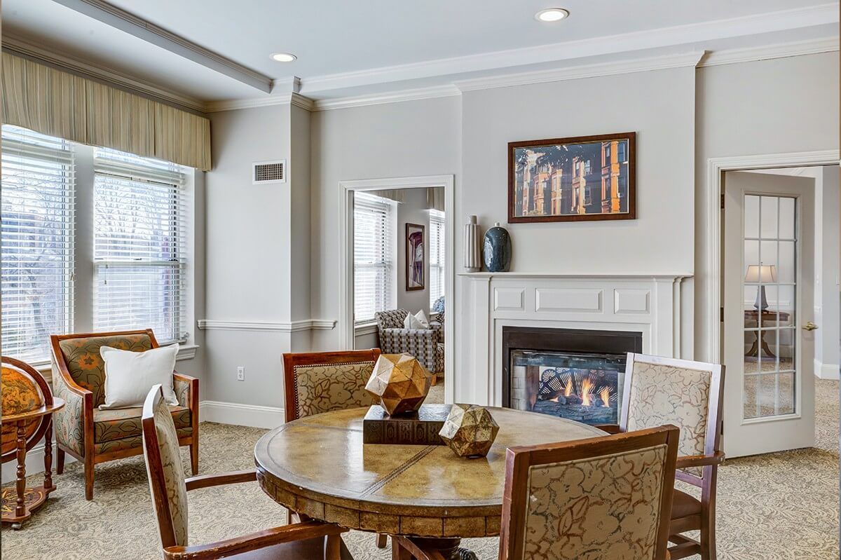 A cozy living room with a round dining table, chairs, and a fireplace, adorned with artwork and large windows.