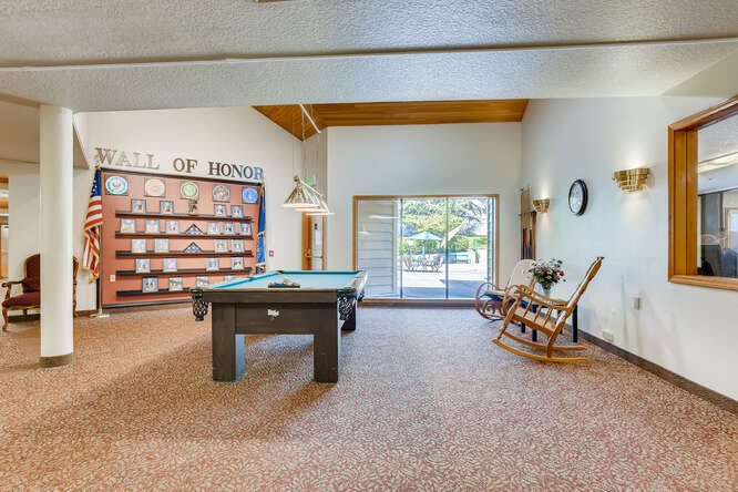 A spacious game room featuring a pool table, bookshelves, and a rocking chair with natural light streaming through a sliding door.