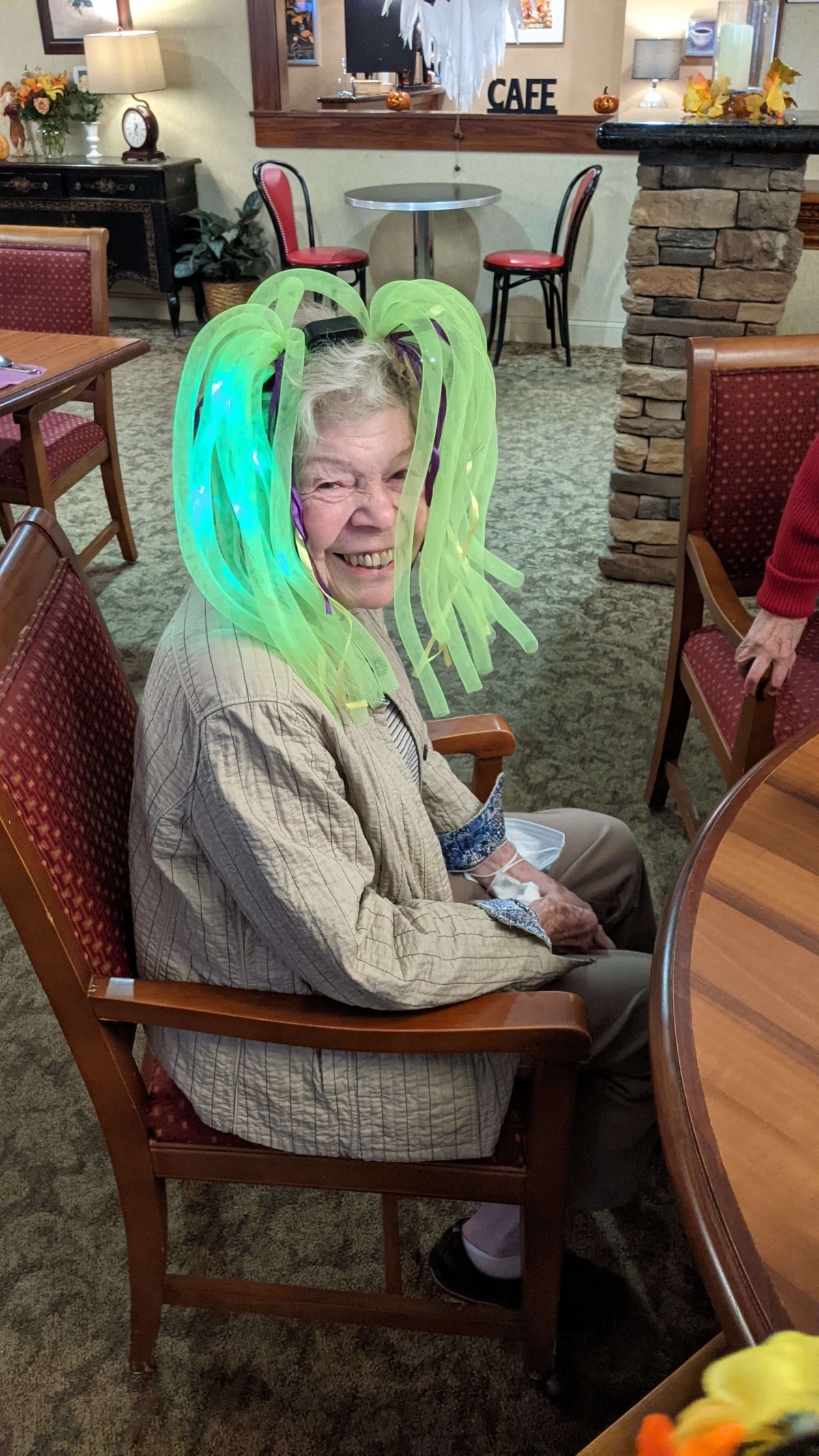 An elderly person wearing a green glowing wig, sitting and smiling.