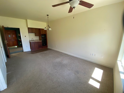 A spacious, empty living room featuring a ceiling fan and access to the kitchen area.