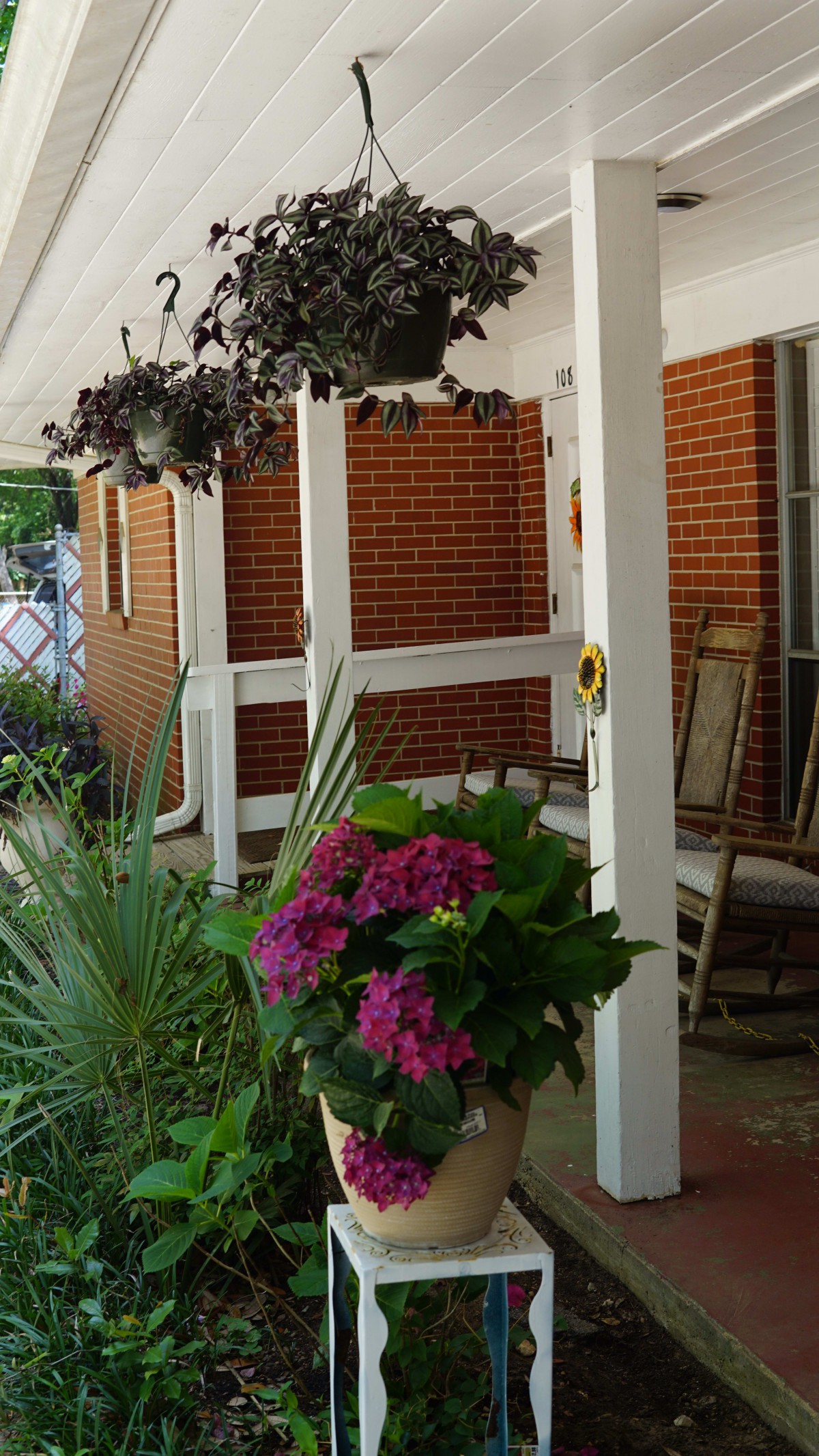 A cozy porch with vibrant plants and flowers, featuring rocking chairs and brick walls.