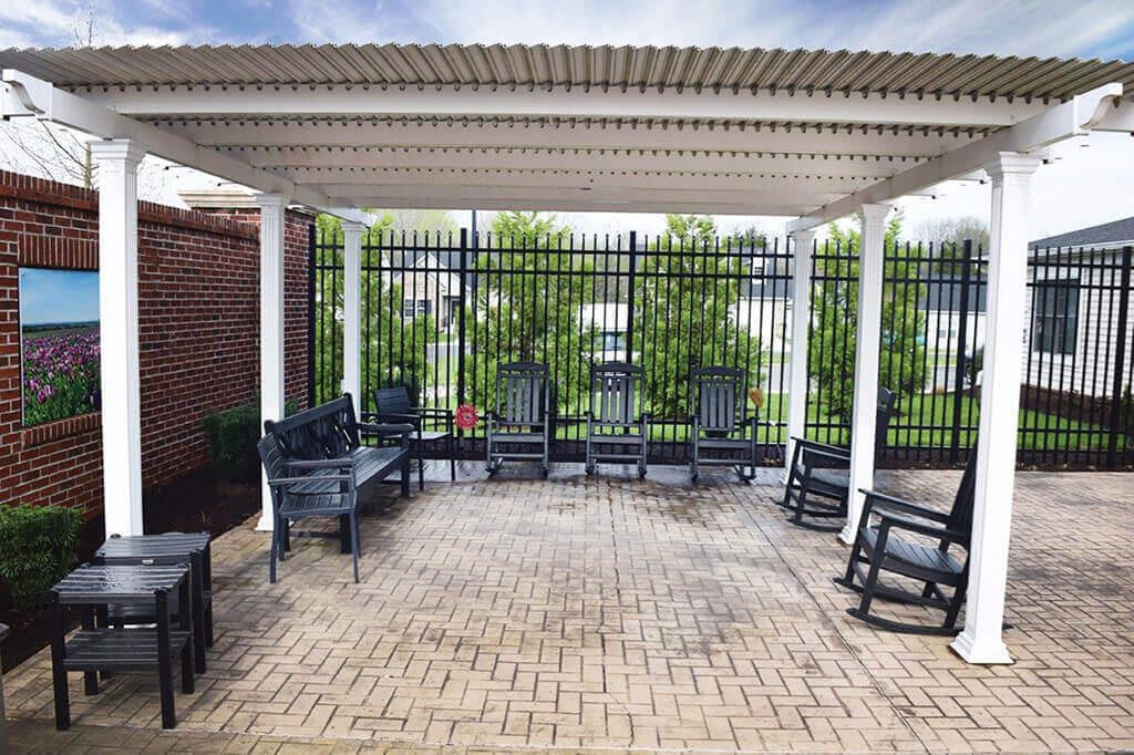 A tranquil outdoor patio with a pergola, featuring rocking chairs and benches on a brick floor.
