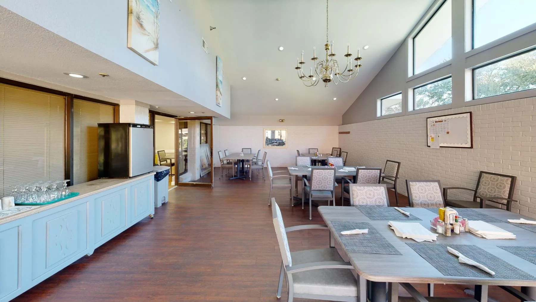 A modern dining area with tables, chairs, and chandeliers, featuring ample natural light from large windows.