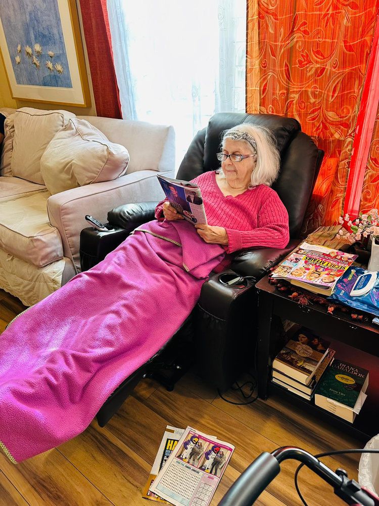 An elderly woman is comfortably reading in a recliner, wrapped in a pink blanket in her cozy living room.
