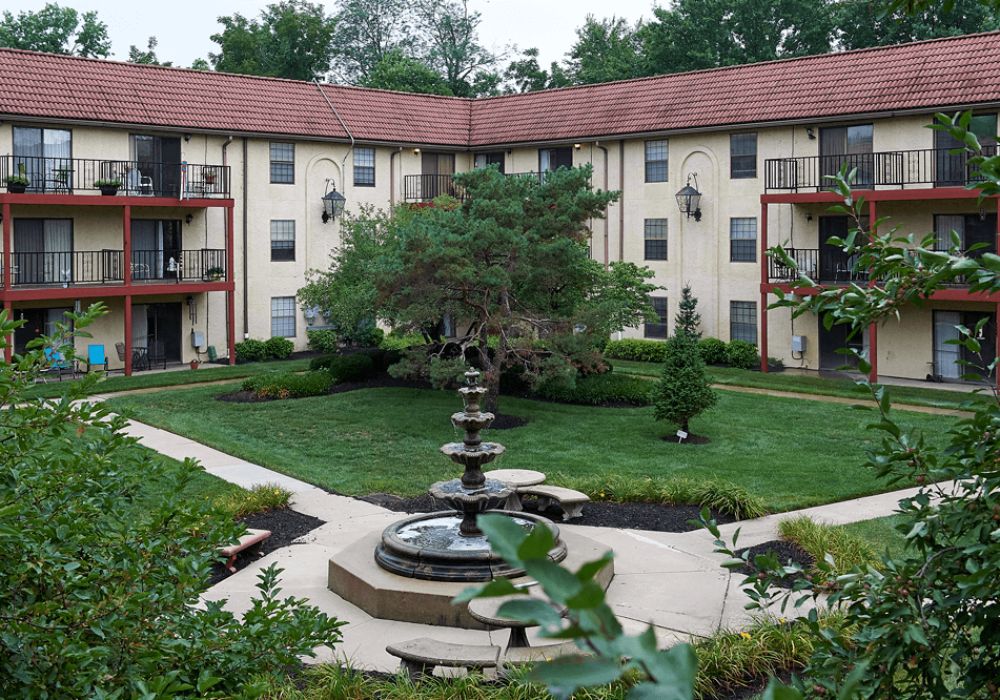A view of an apartment complex with a landscaped courtyard featuring a central fountain and lush greenery.