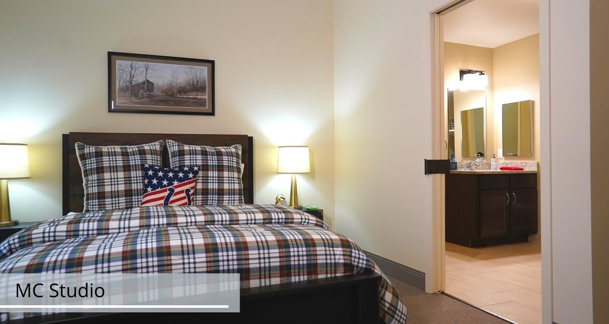 A cozy bedroom with a plaid bedding set, an American flag cushion on the bed, and a lamp on a side table. The room also features wall art and an open doorway.