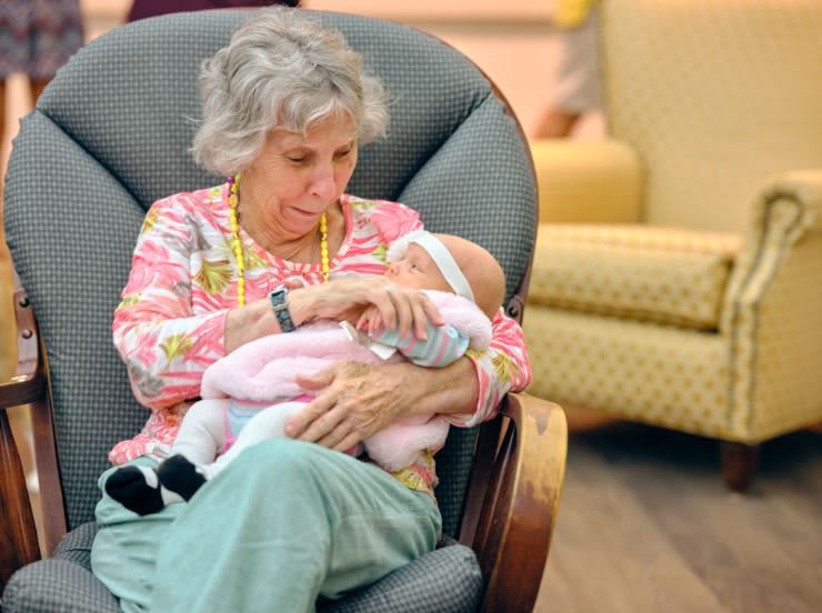 An elderly woman gently cradles a baby in her arms while sitting in a rocking chair indoors.