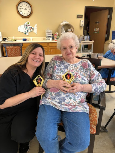 A joyful moment shared between two women as they proudly display their crafts in a senior care facility.