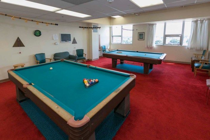 A recreation room featuring two pool tables with a red carpet flooring and large windows.