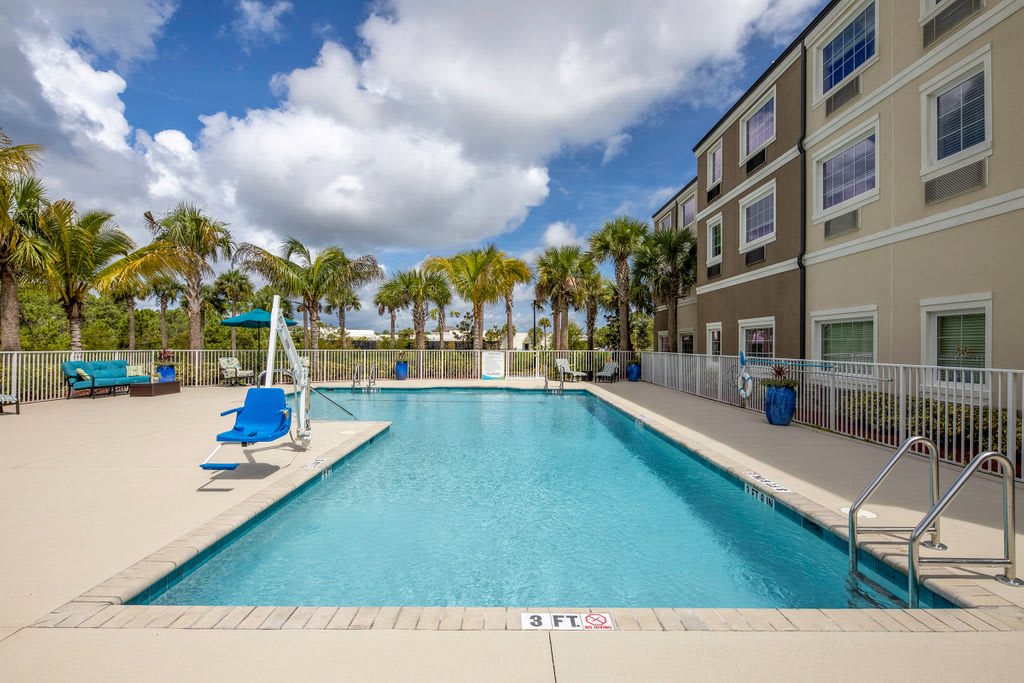 An outdoor swimming pool surrounded by a fence with palm trees and a multi-story building in the background under a cloudy sky.