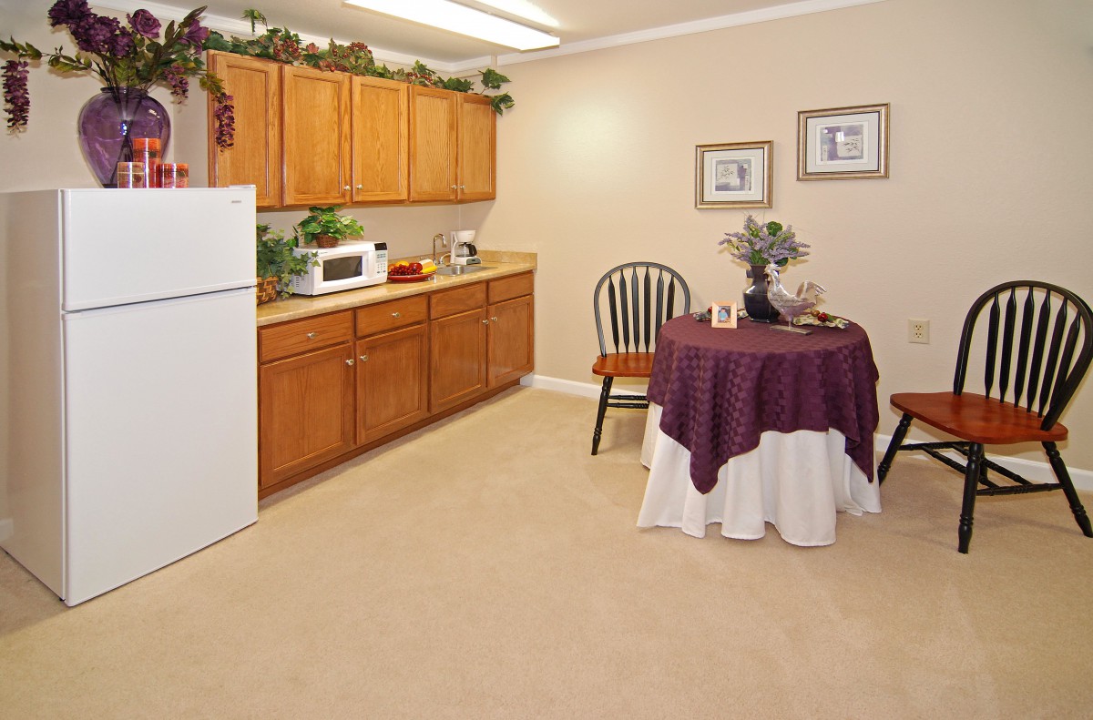 A cozy kitchen and dining area with wooden cabinets, a refrigerator, microwave, and a small round table draped with a purple and white tablecloth.
