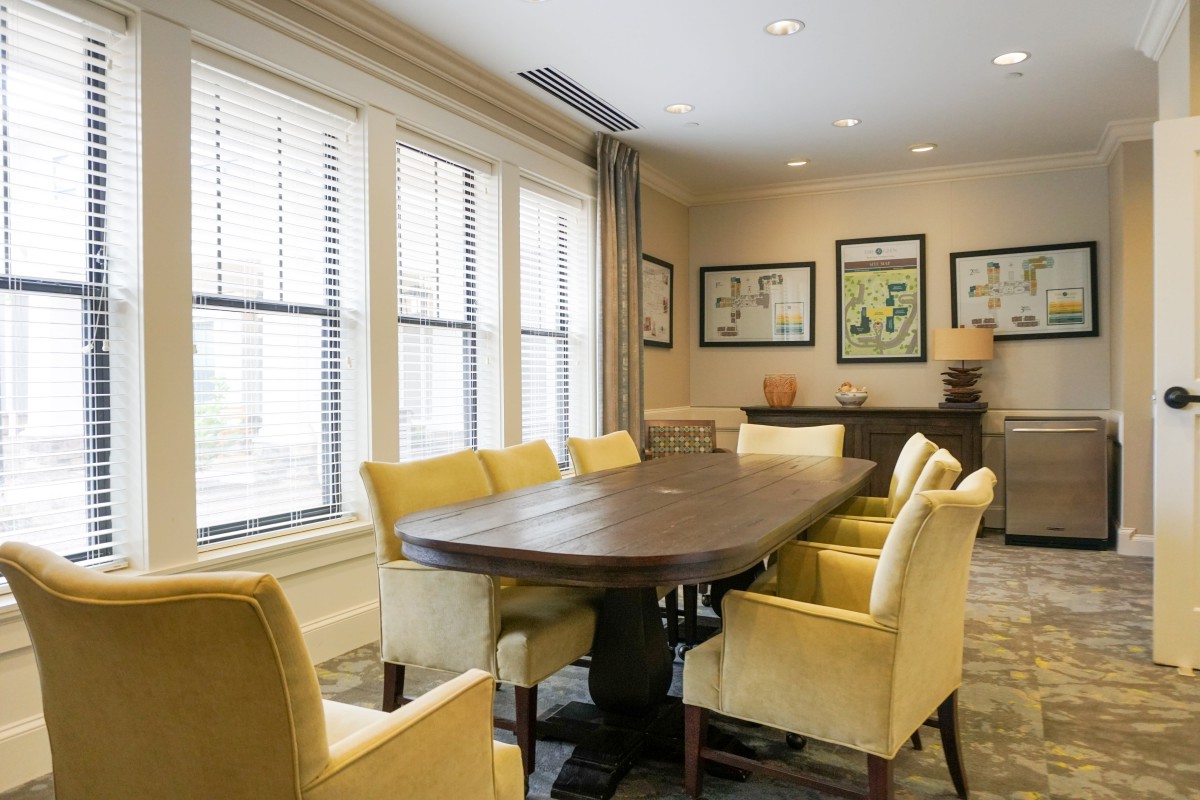 A well-lit conference room featuring a large wooden table surrounded by yellow upholstered chairs, with framed artwork on the walls and natural light streaming through the windows.