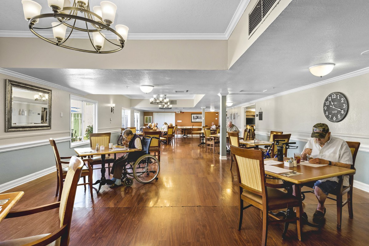 A spacious dining area with wooden flooring, equipped with tables and chairs, featuring a chandelier and a wheelchair for accessibility.