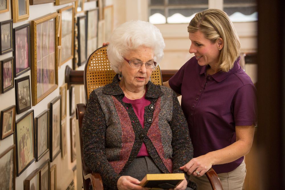 A caregiver comforting an elderly woman holding a book in a home setting.