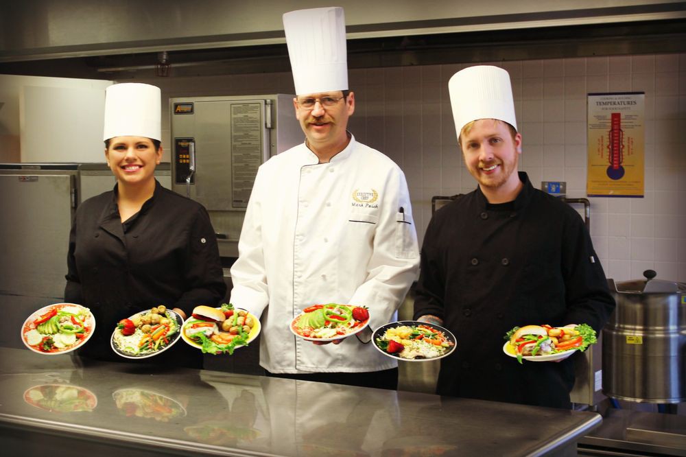 Three chefs proudly display their plated dishes in a professional kitchen.