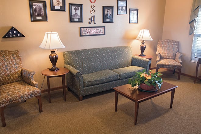 A cozy living room with a sofa, two lamps on side tables, and a coffee table with a floral centerpiece. The walls are decorated with framed pictures.