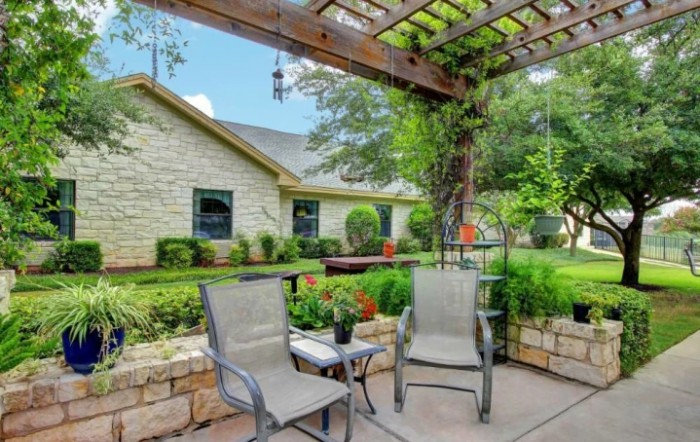 A tranquil patio space with chairs under a pergola, surrounded by lush garden greenery beside a stone house.