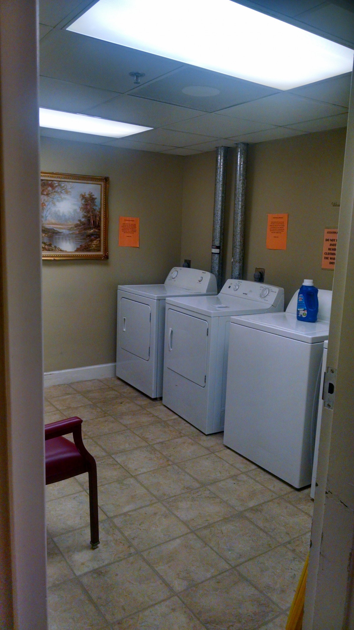 A small laundry room with washing machines and dryers, featuring a bottle of detergent.