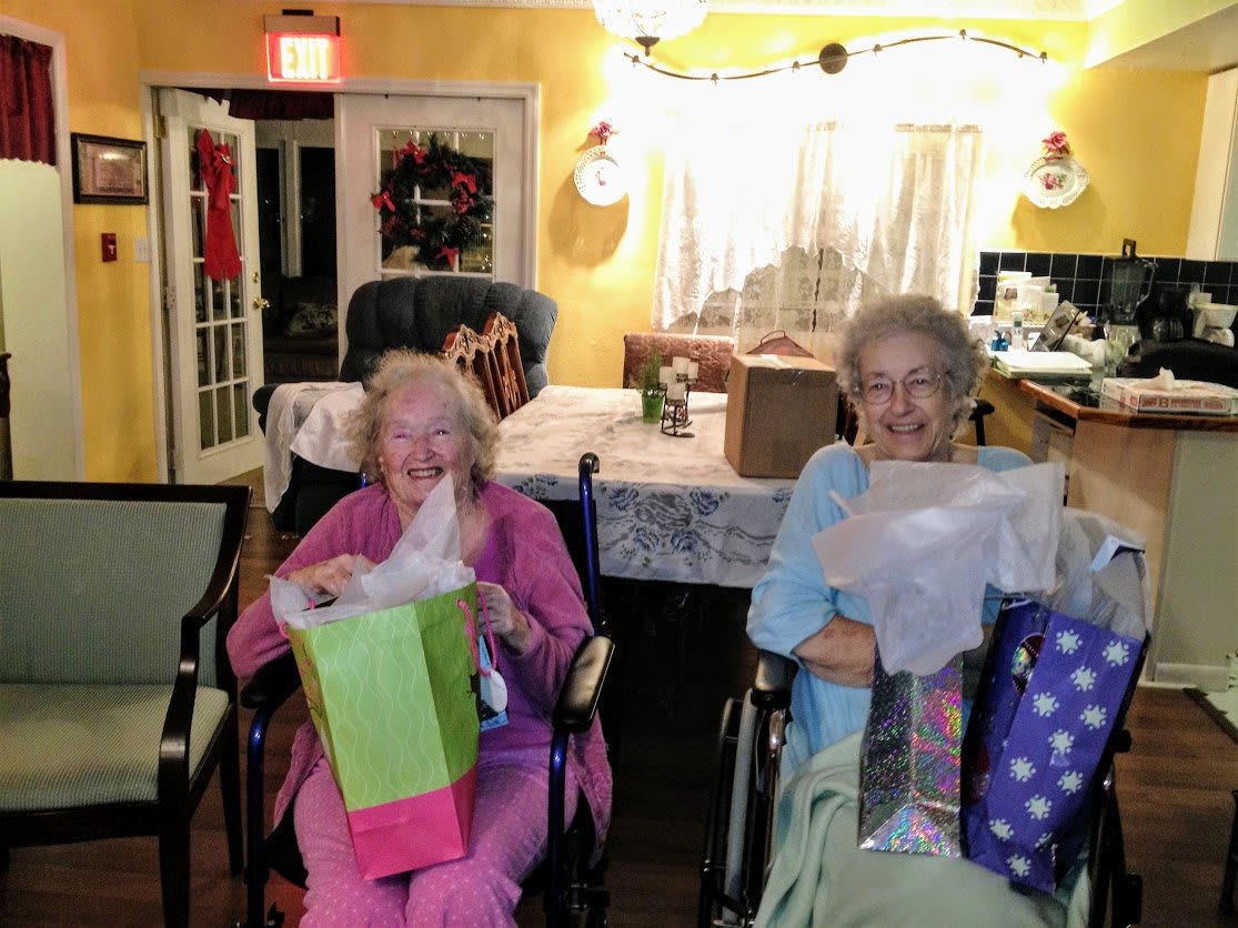 Two elderly women in wheelchairs opening gifts and smiling in a cozy home setting.