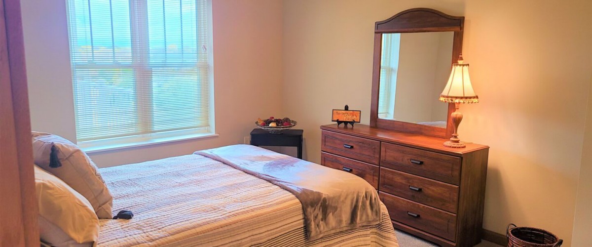 A cozy bedroom with a neatly made bed, wooden dresser, and a mirror by the window.