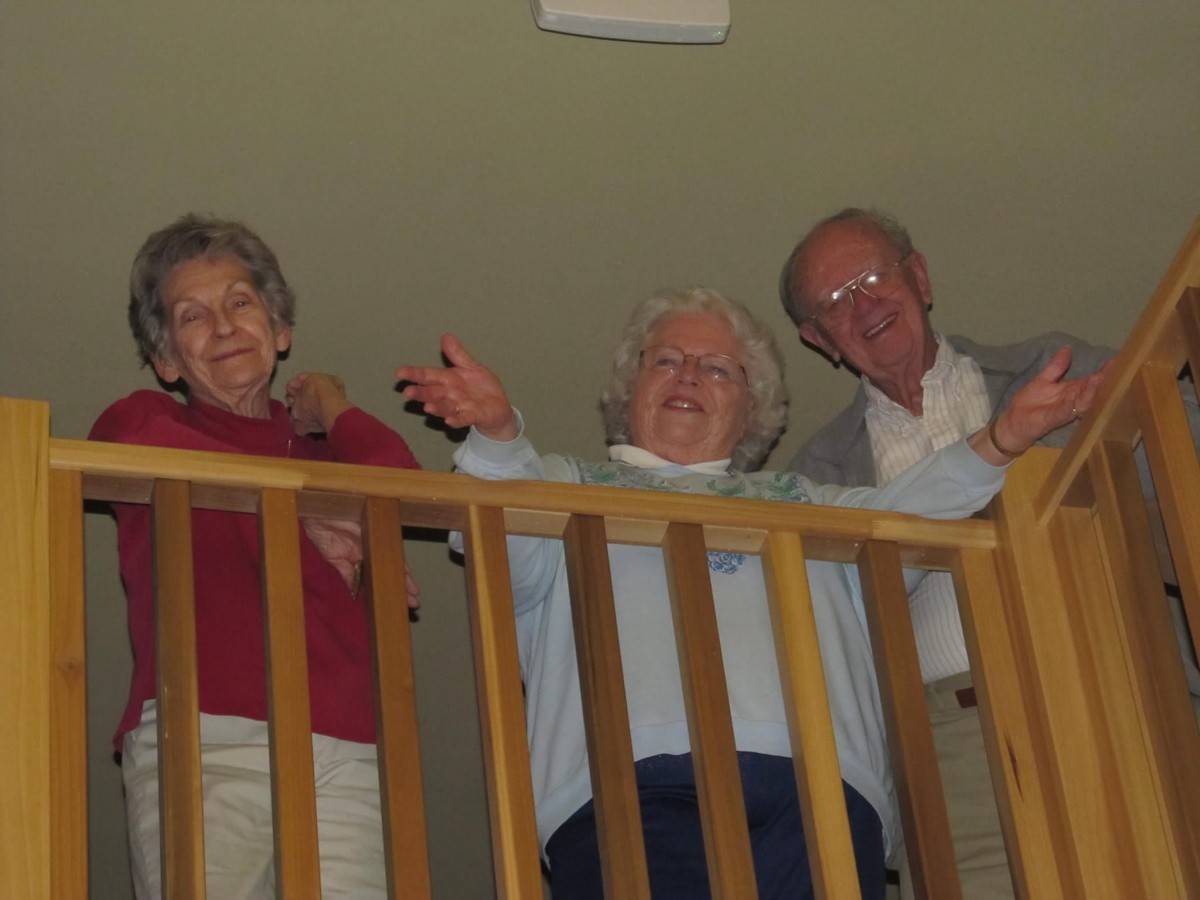Three seniors smiling and waving from a balcony indoors.