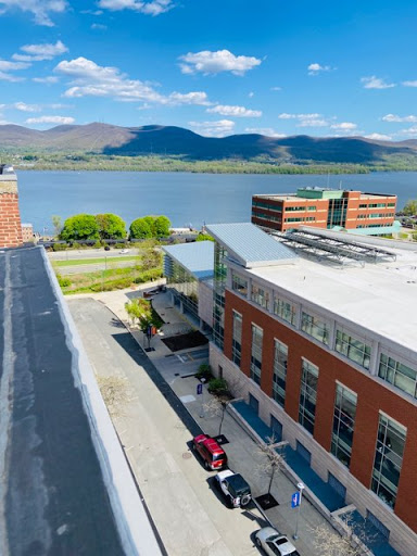 A scenic view of a modern building along the river, framed by mountains under a clear blue sky.