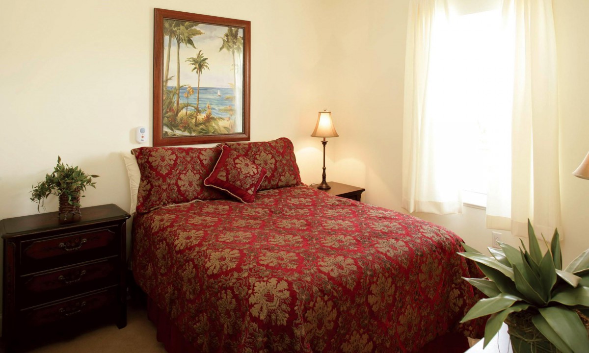 A cozy bedroom featuring a bed with a red floral bedspread, a lamp, and a painting of a tropical scene on the wall.
