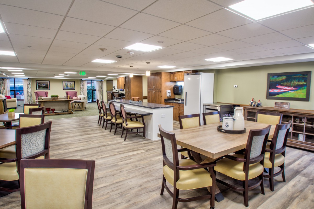 A spacious and well-lit dining area with multiple tables and chairs, featuring a kitchenette in the background.