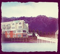 A scenic view of a house by the lake with a pier extending over the water, set against a backdrop of lush forested hills.