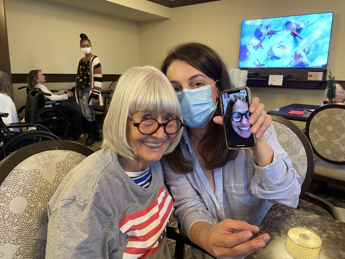 An elderly woman and a younger woman, wearing a mask, happily engage in a video call with another person visible on the phone screen.