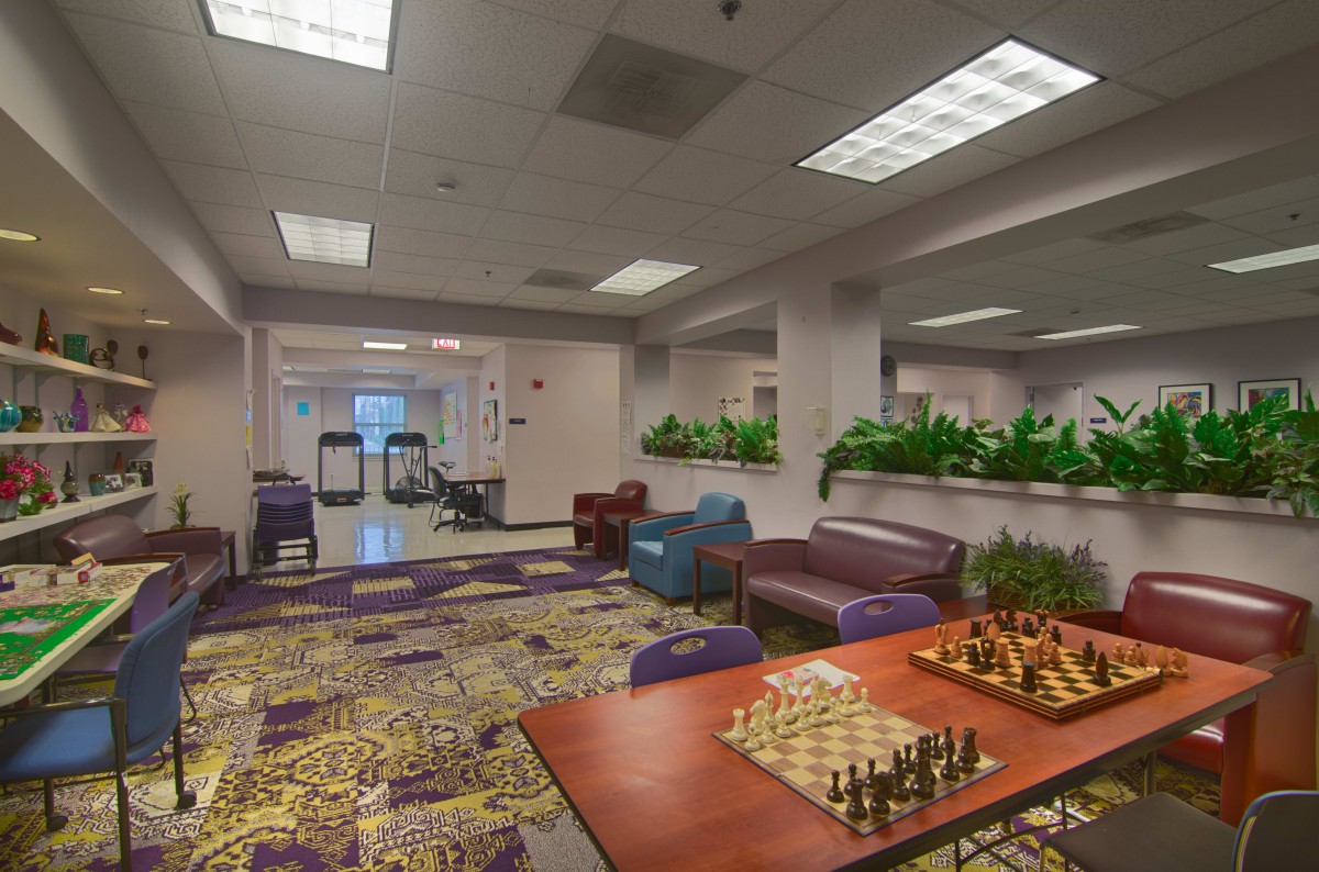 A cozy indoor seating area featuring a chess board on a table, surrounded by comfortable chairs and sofas, with decorative plants and bright lighting.